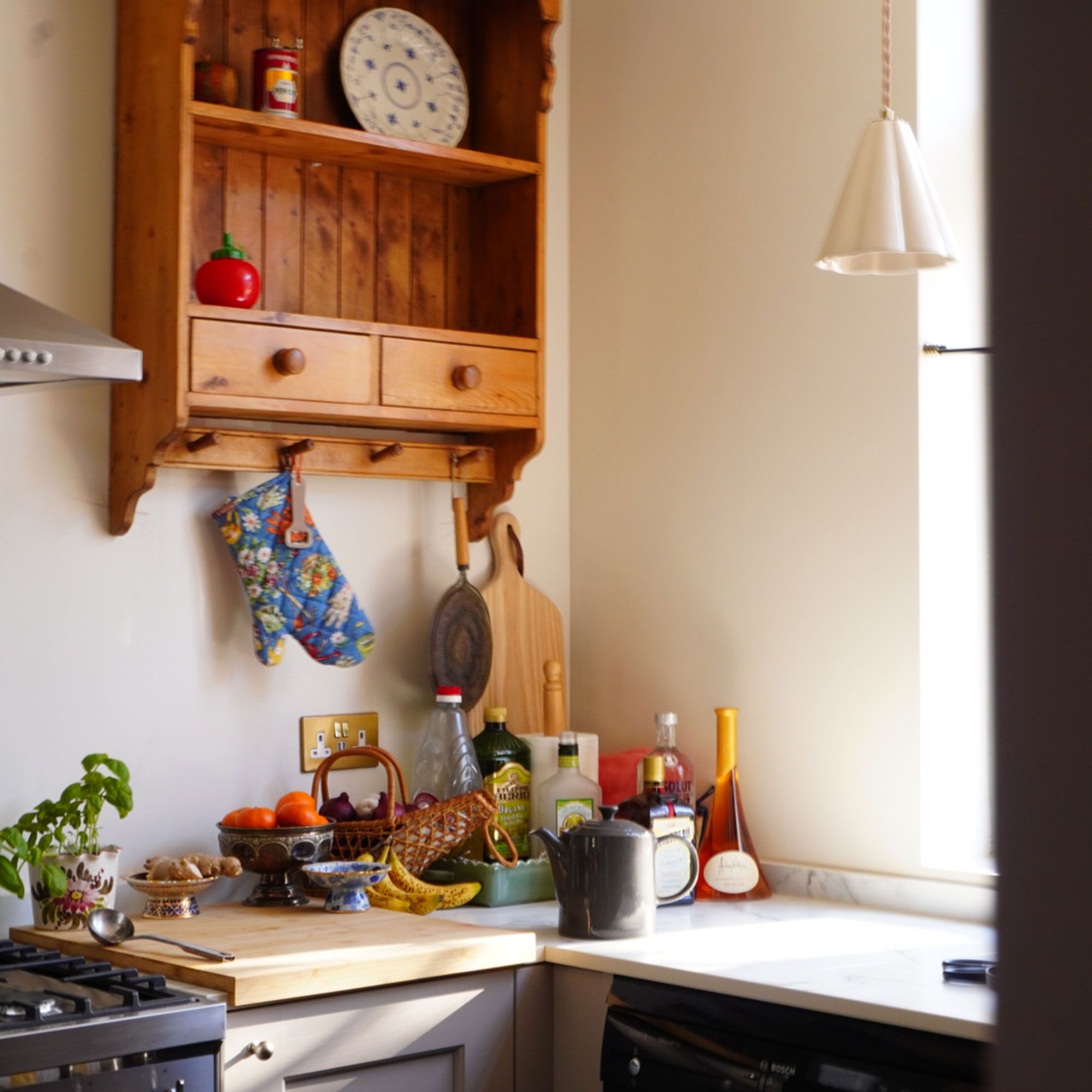 Corner of kitchen with wooden wall storage unit, and collection of bottles, chopping boards and fruit bowls on worktop