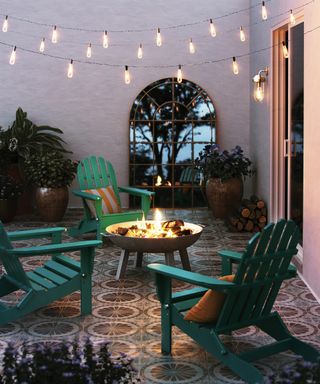 Large round firepit on legs surrounded by green lounge chair, with garden mirror, festoon lighting, and patterned floor tiles.