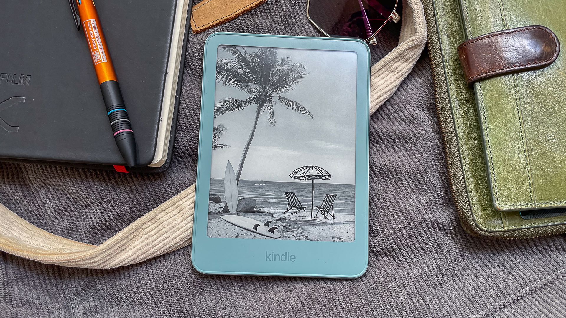 A beachy wallpaper displayed on the Amazon Kindle 2026 while it lies on a cloth bag and surrounded by handbag contents