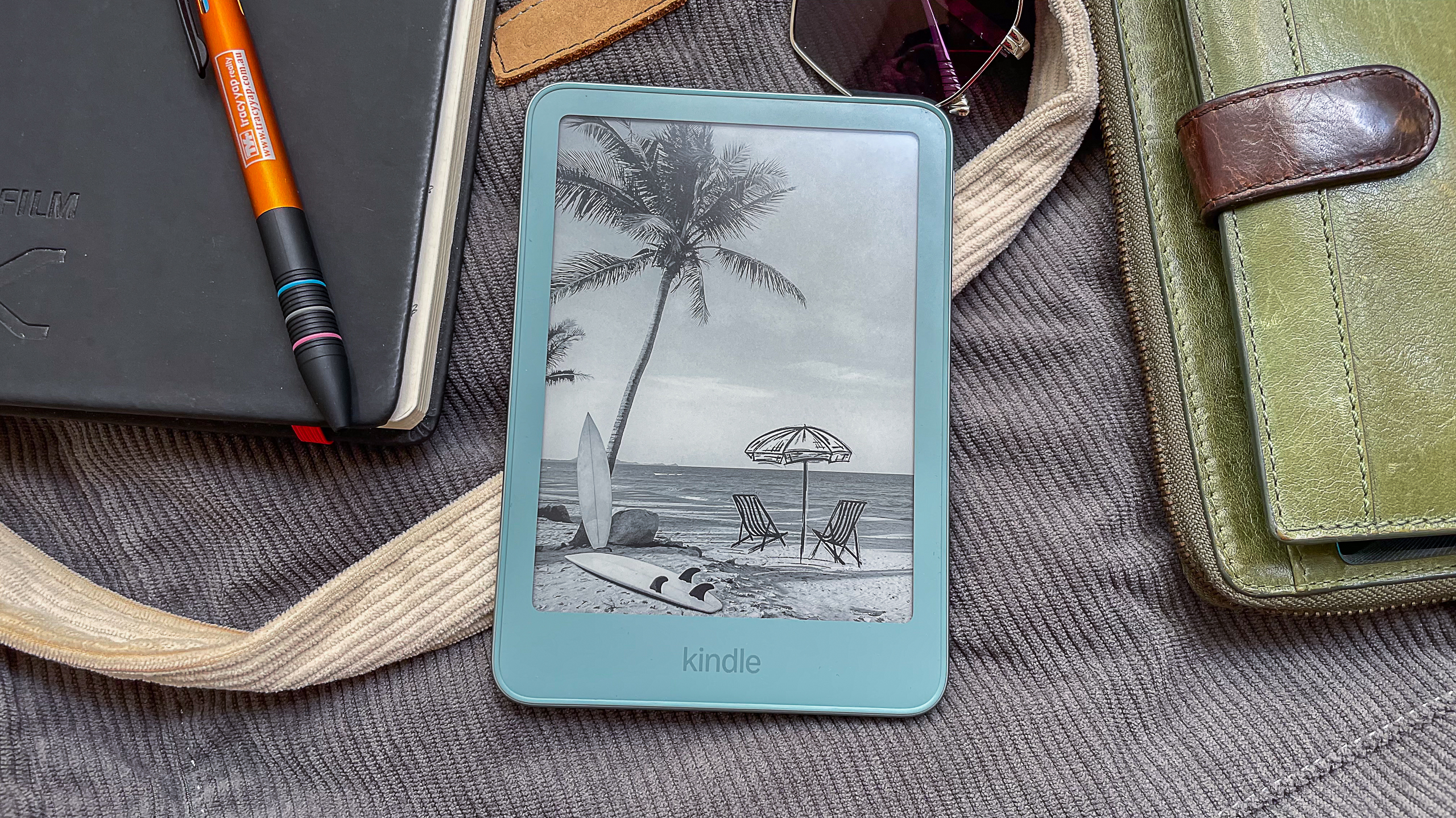 A beachy wallpaper displayed on the Amazon Kindle 2026 while it lies on a cloth bag and surrounded by handbag contents