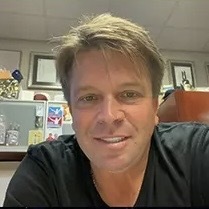 A middle aged man in a t-shirt is smiling from the chair of an office room.