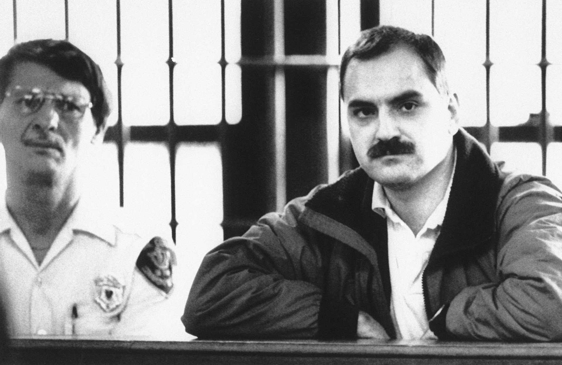 a mustachioed man frowns while being booked into a prison