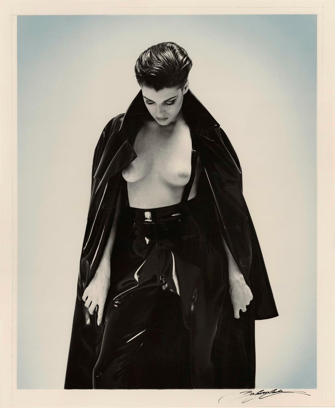A black and white photograph of a nude woman reclining on a leather sofa in high heels, holding a lit cigarette.