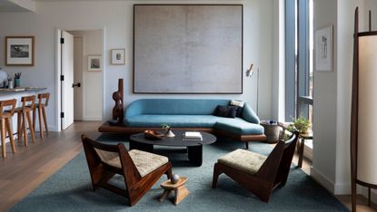 A living room with white walls, dark wood flooring, muted blue rug and brighter blue sofa, with large grey wall art and dark wood armchairs