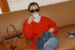 Model wearing white t-shirt, jeans, and red Almina Concept v-neck sweater, sitting on couch and drinking out of coffee cup.