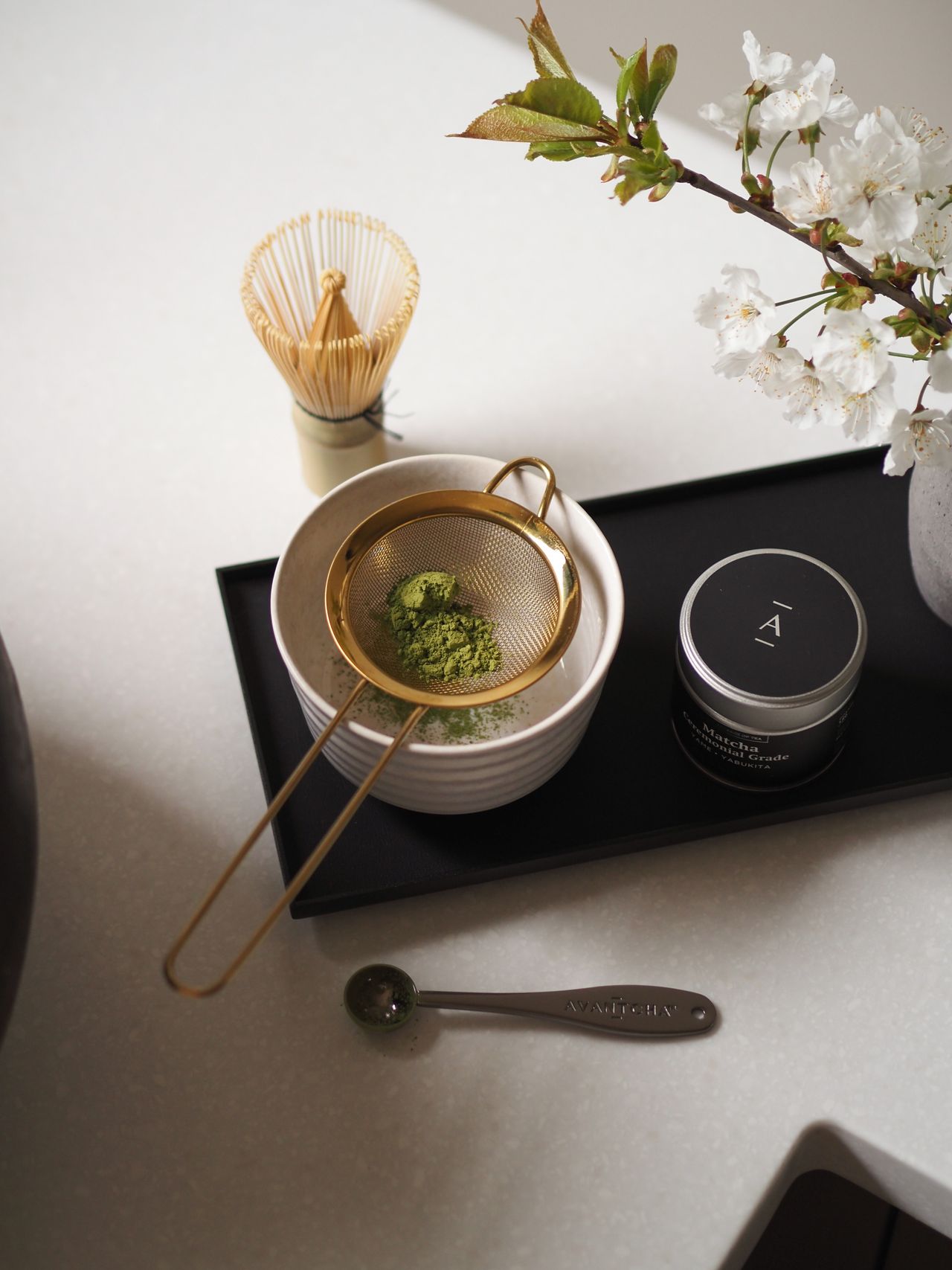 A matcha tea tray photographed from above in a naturally lit room, placed on a black tray next to a wicker wisk on a white countertop decorated with a vase of fresh cherry blossom.
