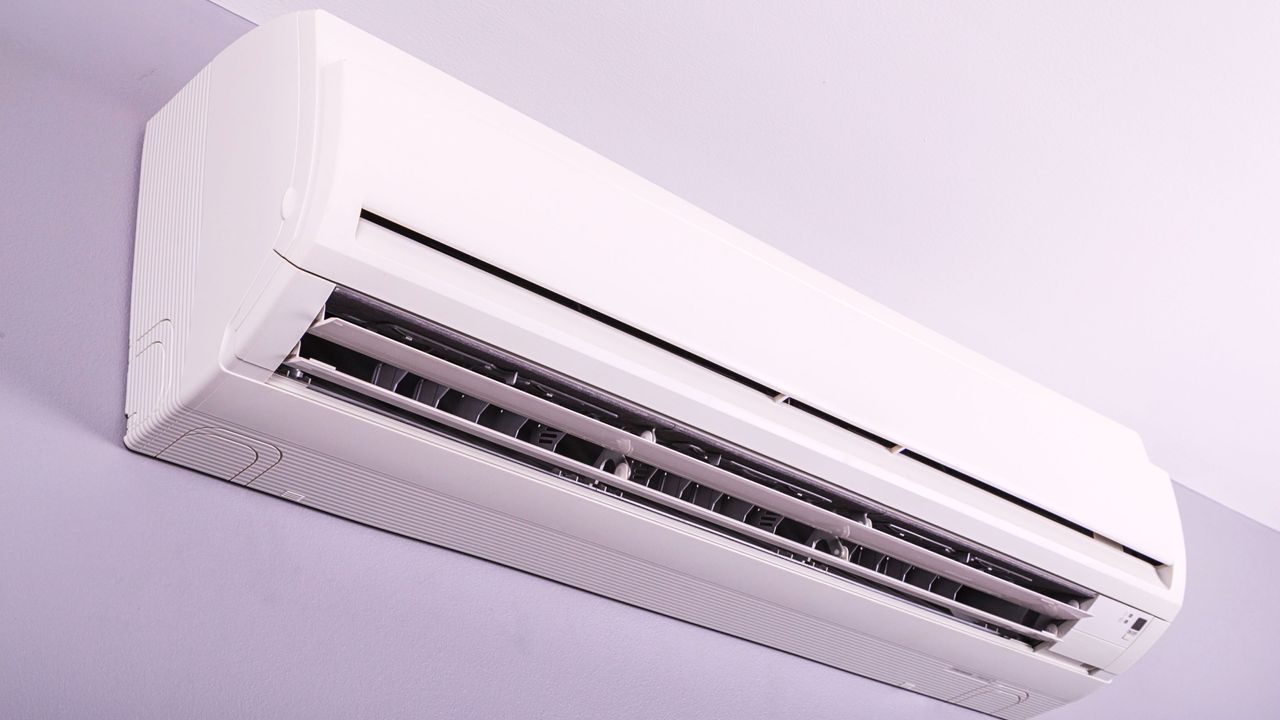 We answer &#039;how long does it take for an AC to cool a room?&#039; Here is a white rectangular AC unit with a black vent, mounted onto a light purple wall