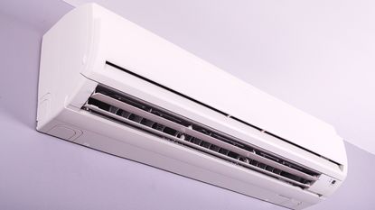 We answer 'how long does it take for an AC to cool a room?' Here is a white rectangular AC unit with a black vent, mounted onto a light purple wall
