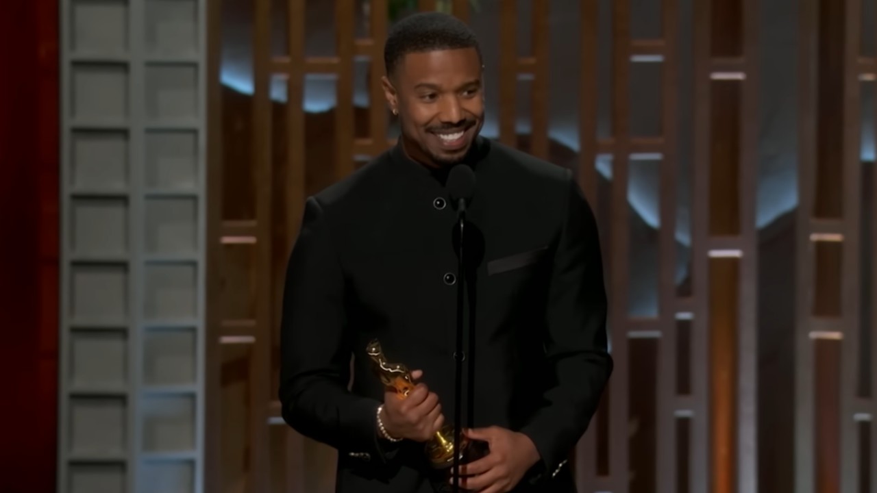 Michael B. Jordan smiling while talking into a mic and holding his Oscar. 