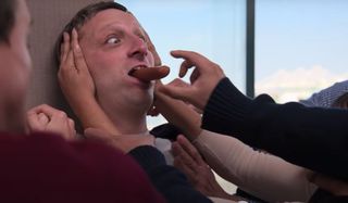 Tim Robinson is surrounded by a crowd trying to snag a hot dog from his mouth in I Think You Should Leave.