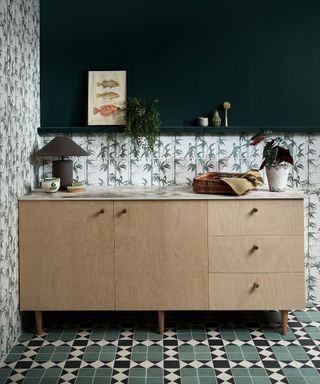 A room with a light wooden console table with lamps and flowers on it, a dark green wall behind it, and dark green and white tiles around it