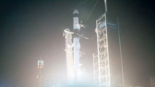 a black and white rocket ignites its nine engines, lighting up its launch pad in the darkness of night