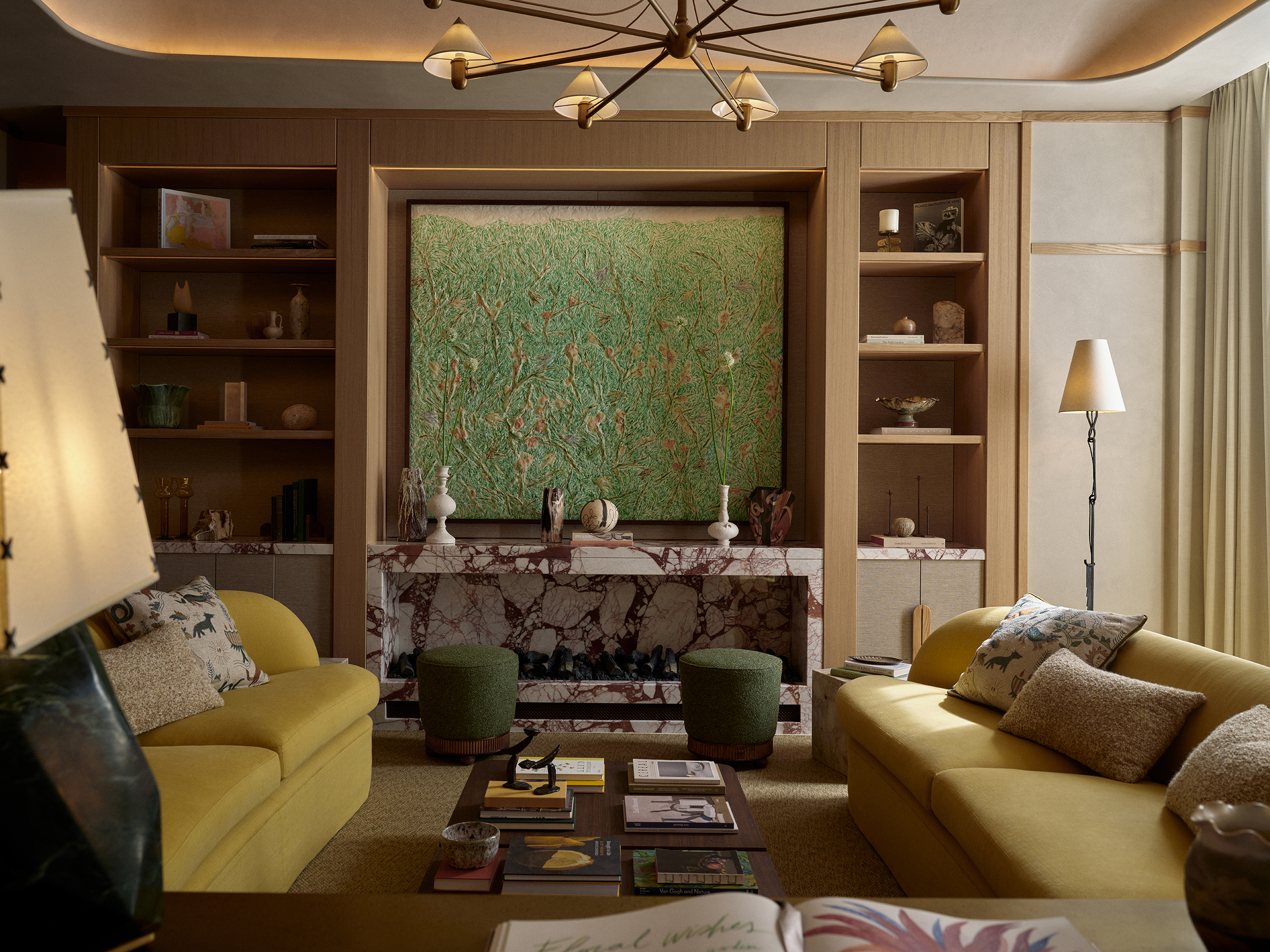 A warmly decorated hotel lobby featuring oversized couches in ochre, floral-inspired artwork, chocolatey wooden tones, a curated selection of coffee table books, floor lamps with softly glowing paper shades, and more.