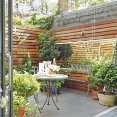 small enclosed garden with wooden cladding walls, large rectangular mirror, containers and pots, crittall doors, iron bench, table and chairs 
