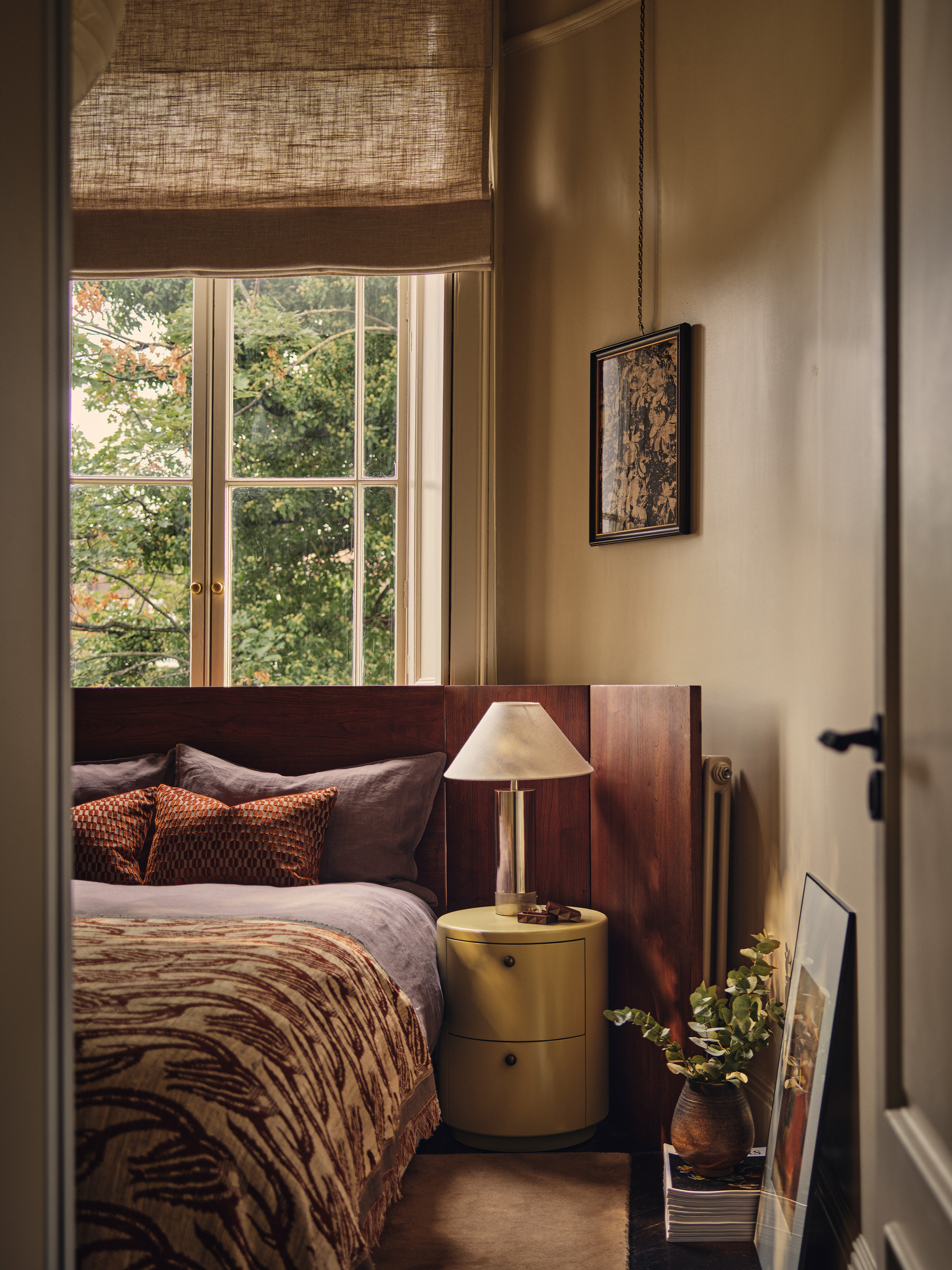 Image of an off-white bedroom with a large bed that has an extended wooden headboard. The bedding is purple and red, and there is a lamp by the bed.