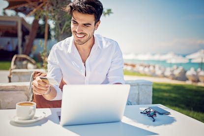 A bearded man holds a gold credit card and orders on a computer at an ocean-side cafe.