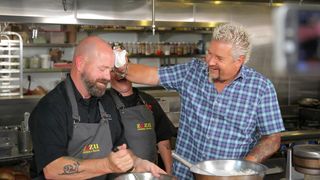 (R to L) Guy Fieri pats down the forehead of a cook in Diners, Drive-Ins, and Dives
