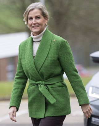 Duchess Sophie wearing a green blazer with a tie waist and a tan turtleneck walking through a parking lot and smiling