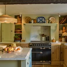 country kitchen with rattan pendant lamp above a sage island with marble worktop surrounded by oak cabinetry with black range cooker 