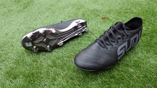 A pair of Black football boots, one with a silver underside and the other with 'SUDU' written on the side lay next to eachother on green artificial grass. Black Sudu SFS FG 01+ Pro