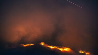 orange flames glowing from a black pit of clouds reflect off smoke in the sky as a shooting star streaks by the upper right.