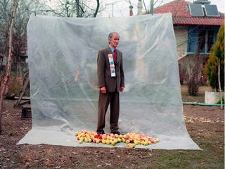 An grey-haired man wearing a brown suit is stood against plastic sheeting with dozens of apples at his feet in an outside setting.