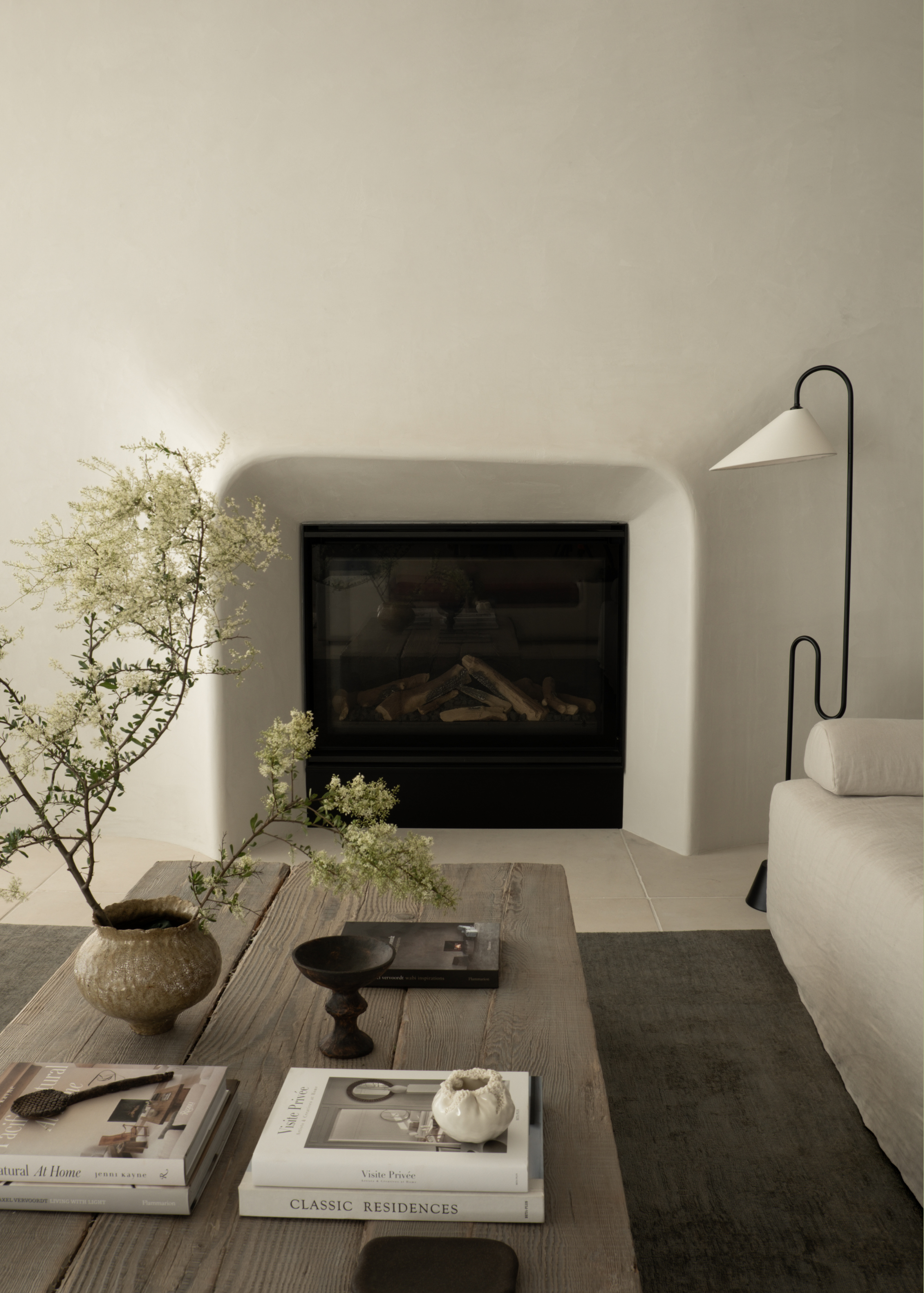 A living room with white walls, a fireplace, a curved floor lamp, a low wood coffee table with books and a vase with stems