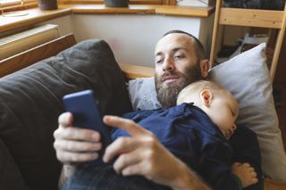 Dad lying on the sofa looking at his phone with his infant son asleep on his chest