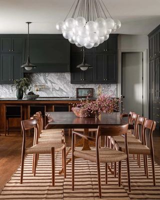 shot of kitchen and dining room space with a wooden dining table and 10 wooden chairs around it. There is black cabinetry and white marble backsplash in the kitchen. A brown and beige striped rug is under the table