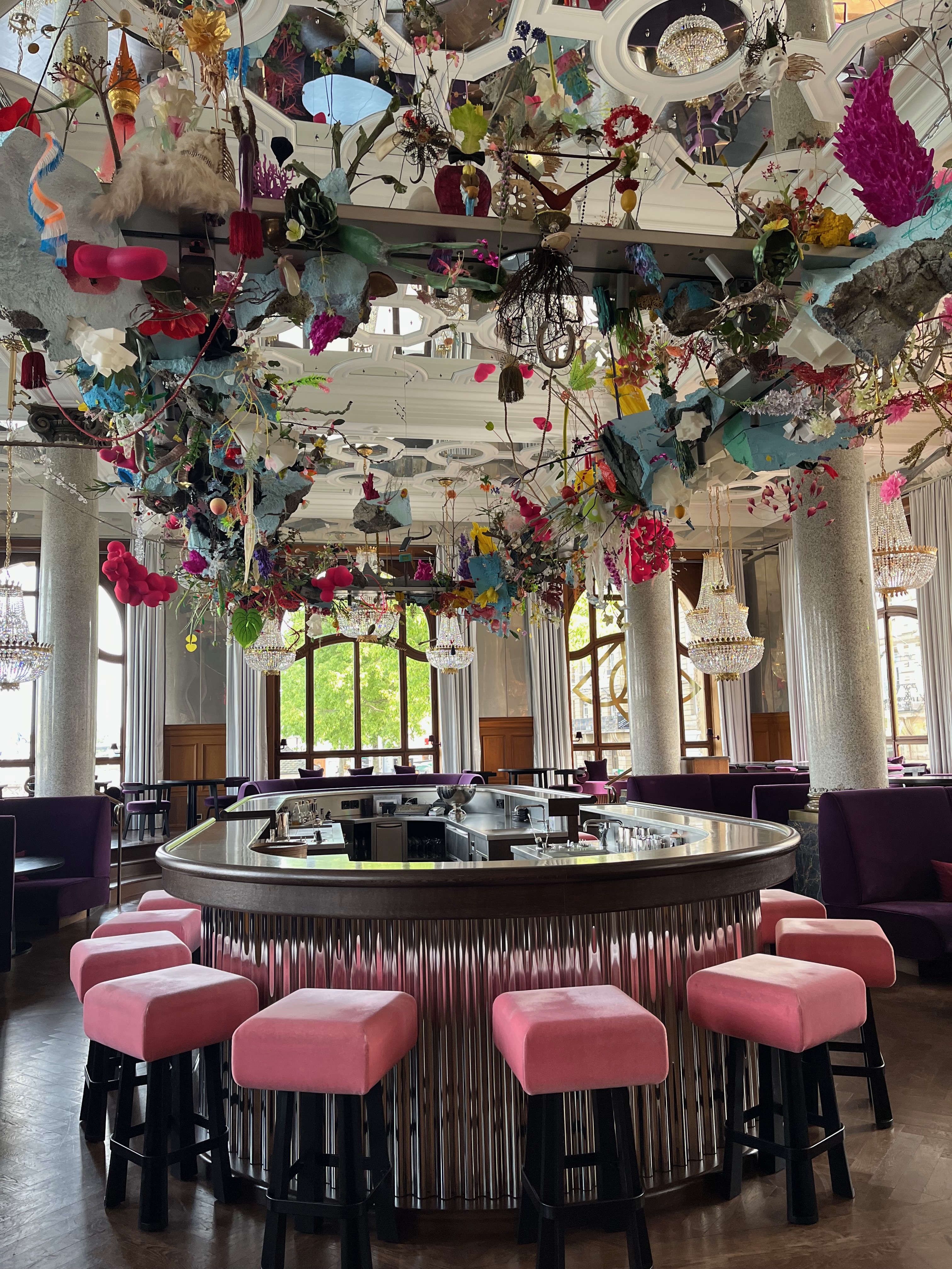 The interior of Banks, a bar inside Les Trois Rois, featuring pink velvet barstools with black legs, a silver bar, and multi-colored decorations hung from the ceiling.