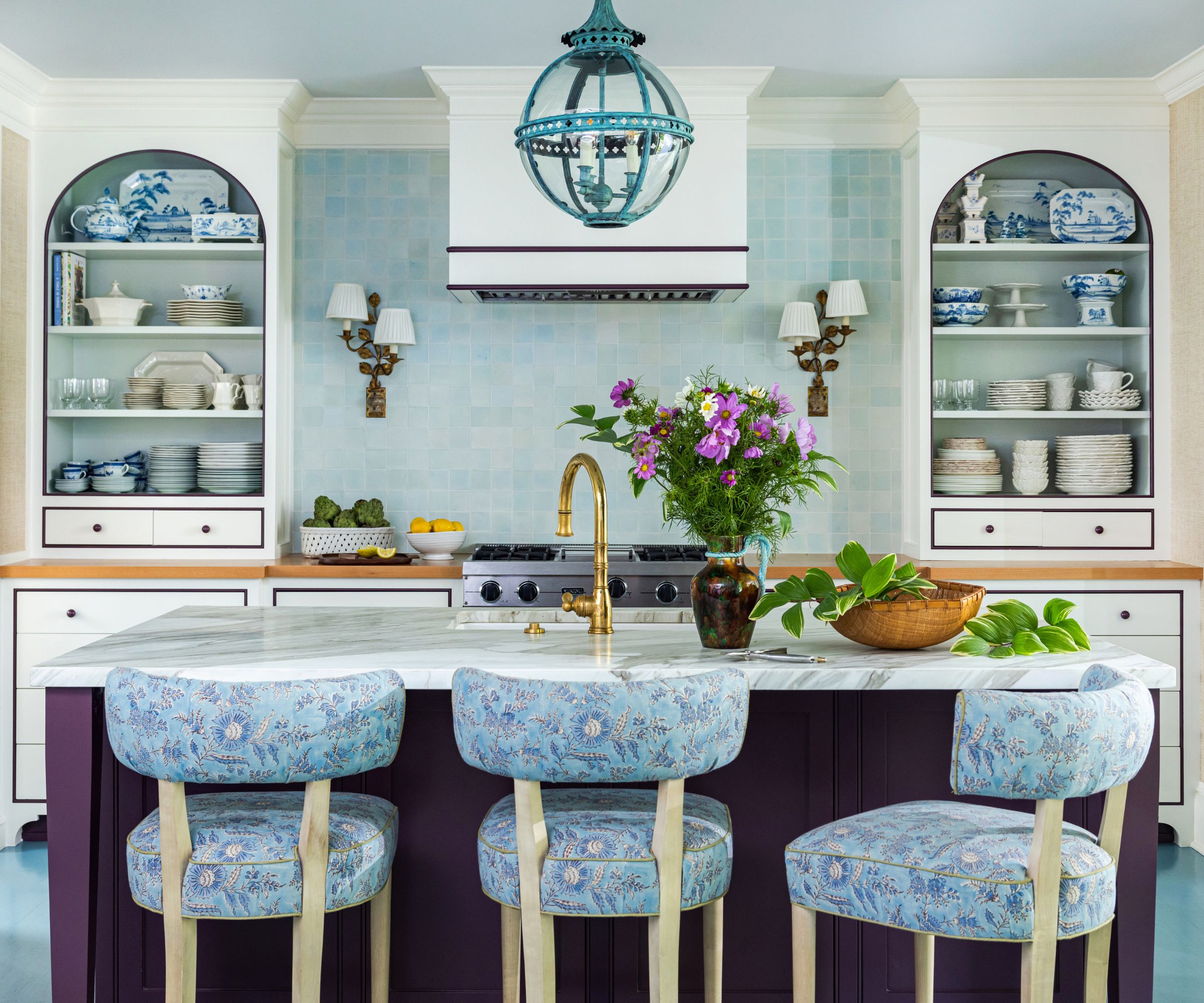 Kitchen with pale blue tiled backsplash, open shelves with blue and white china, an aubergine painted island with a marble top and upholstered stools in a floral blue fabric