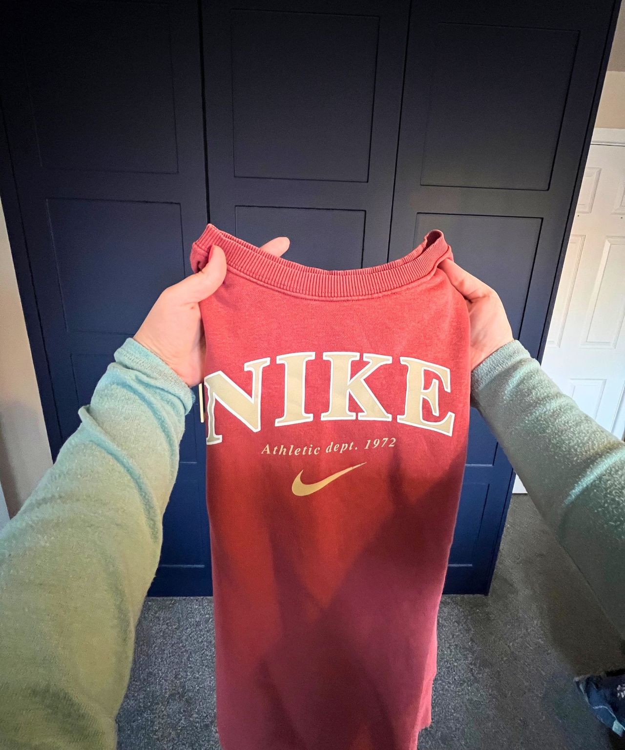 The image shows a point of view shot of a peachy-orange Nike long sleeve sweater being held in front of the camera with the arms now folded back in the air