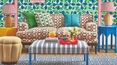 A bright patterned living room with patterned wallpaper, couch, footrest, and rug. The floor is jute carpet, and to either side of the sofa are matching rattan lamps on mismatched tables.
