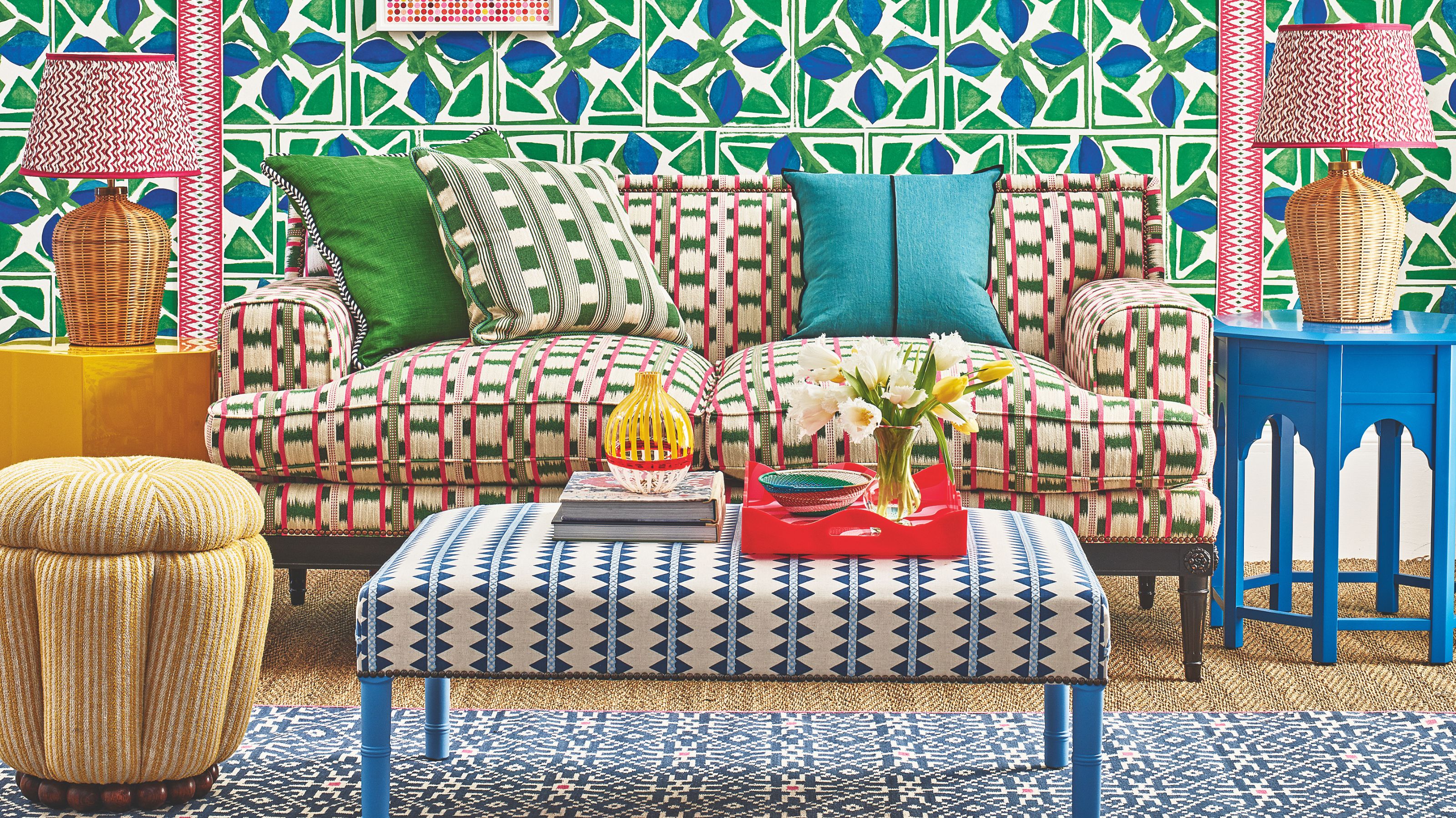 A bright patterned living room with patterned wallpaper, couch, footrest, and rug. The floor is jute carpet, and to either side of the sofa are matching rattan lamps on mismatched tables.
