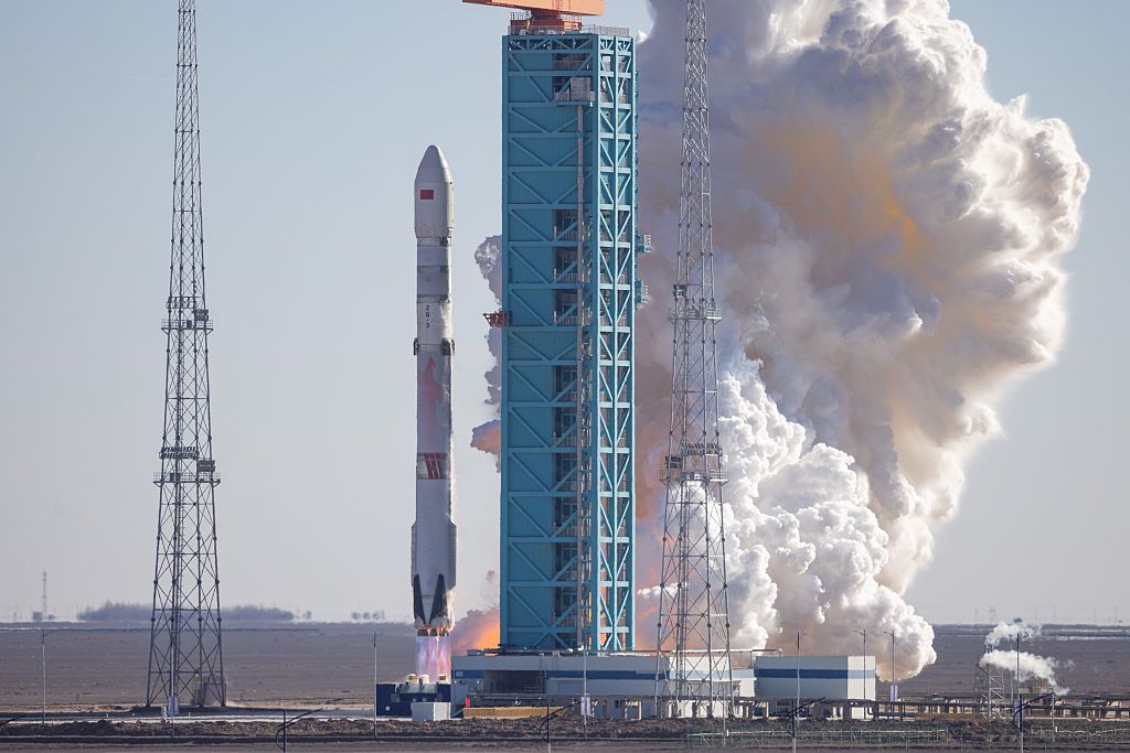 A tall white rocket launches from a pad with white smoke in the background