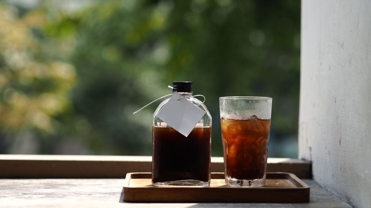 cold brew vs iced coffee, a bottle of cold brew next to a glass of iced coffee on a board in front of a blurred window showing greenery