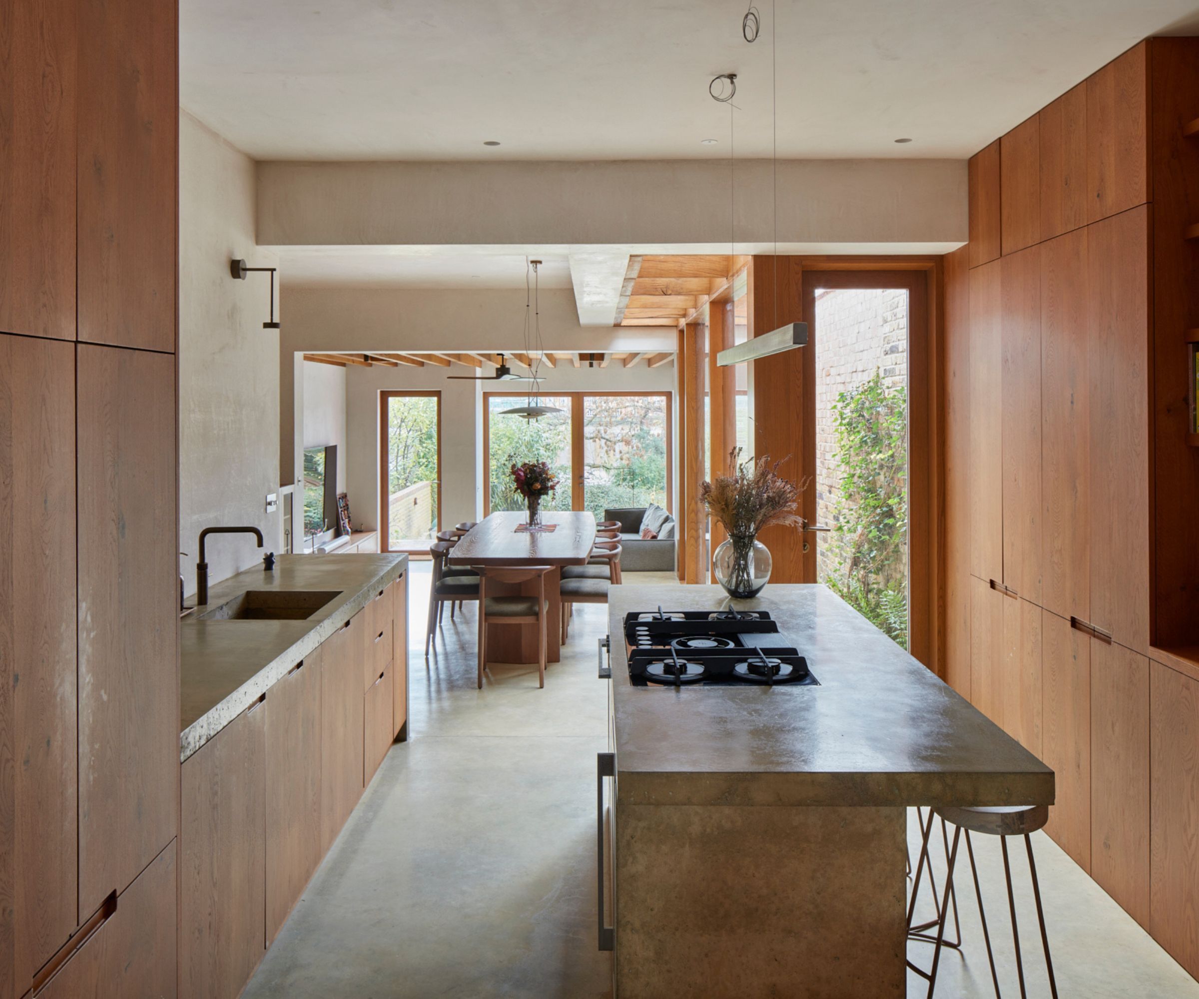 Open-plan kitchen with warm wood cabinetry, concrete island and worktops, and a long dining table aligned with large garden-facing windows.