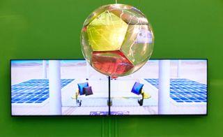 A large scale spherical chandelier made of hexagonal coloured shapes hangs against a bright green wall displaying a widescreen digital image.