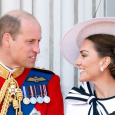 Prince William wearing a red military uniform with medals turning to look at Kate Middleton who is wearing a big white hat and white dress with a navy bow and smiling