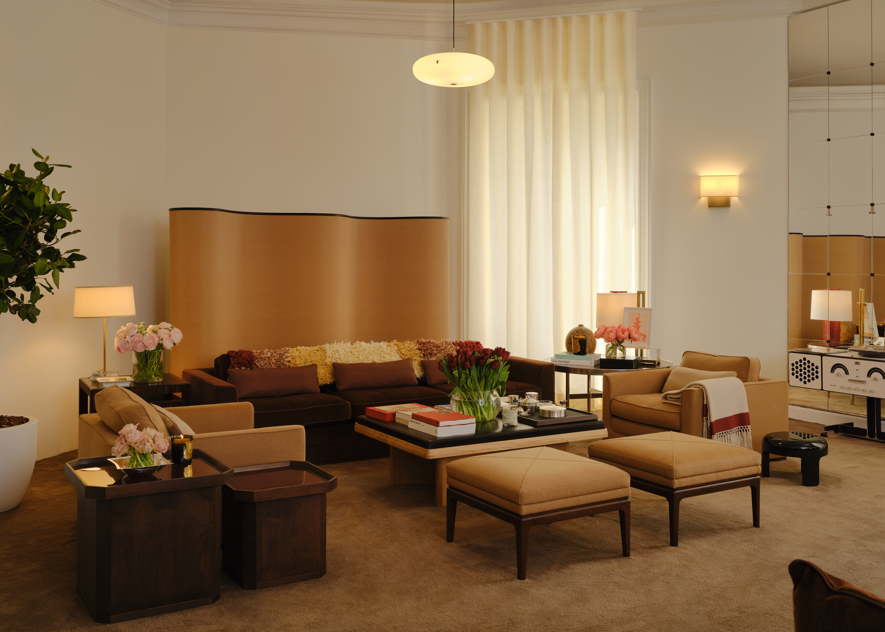 Interior shots of a beige and brown-tinted design atelier decorated with 1970s-inspired furniture designs, pendant lights, collectibles, and plants, and lots of preparatory sketches.