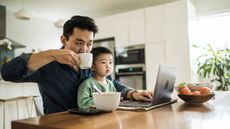 A man sips coffee as he works from the kitchen table with his little boy on his lap.