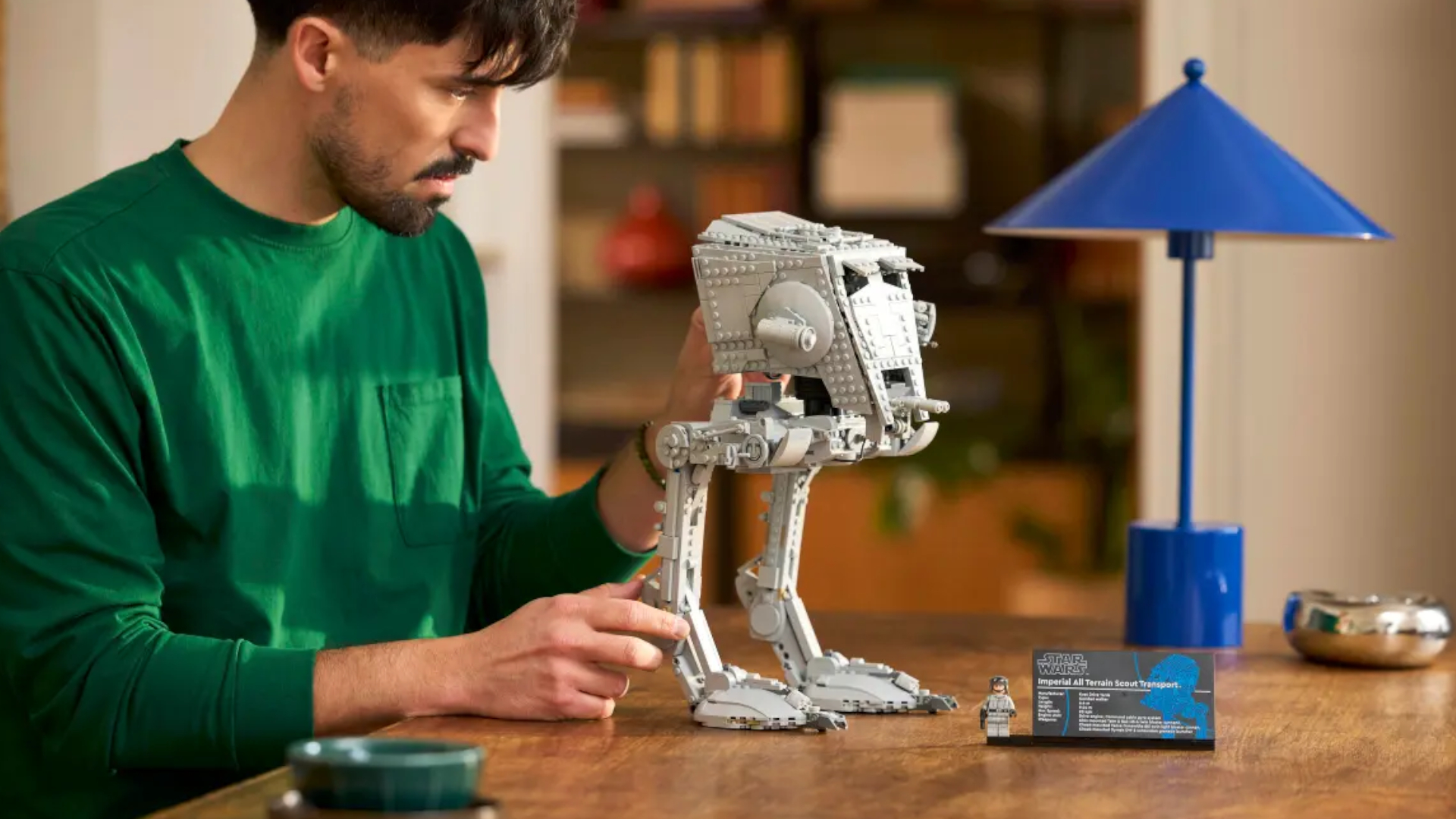A man moves the Lego AT-ST Walker as it stands on a wooden surface beside a plaque and blue lamp