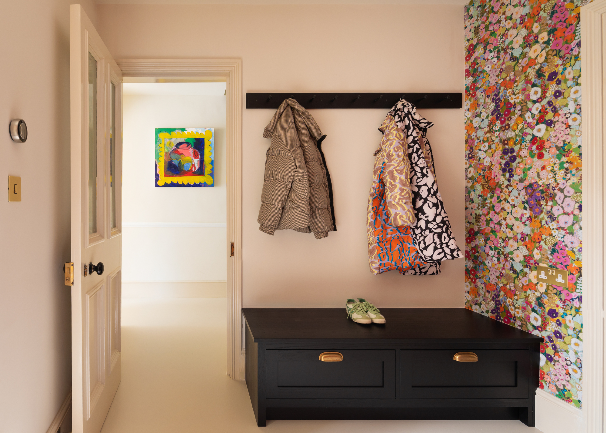 A bright white entryway with a floral-wallpapered accent wall by a black shoe storage bench below a row of hooks