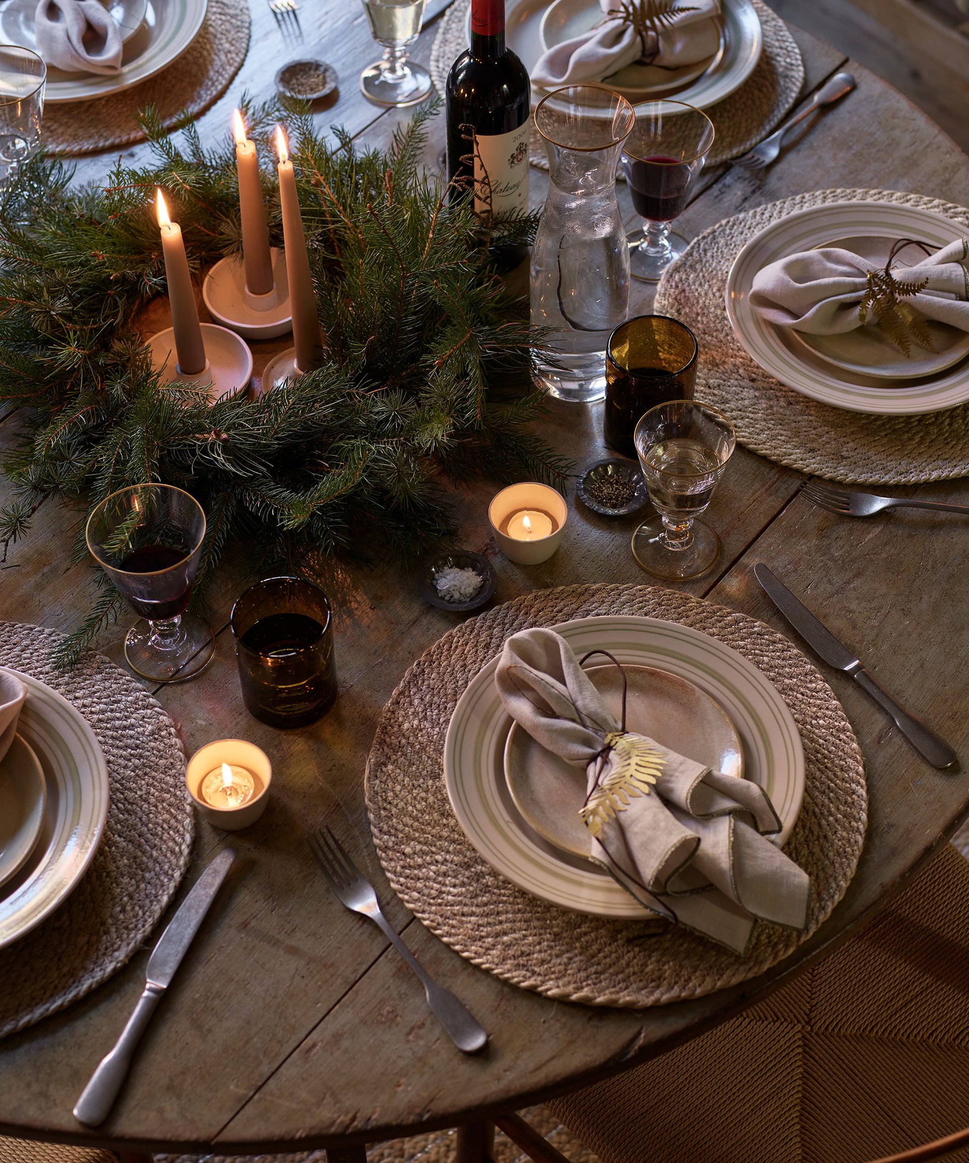 A round wooden table set with woven placemats, white plates, and candles in the middle of a green wreath.