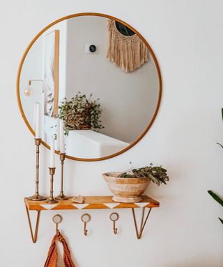 An entryway with a circular mirror, a wooden shelf with candles on, and gold hooks