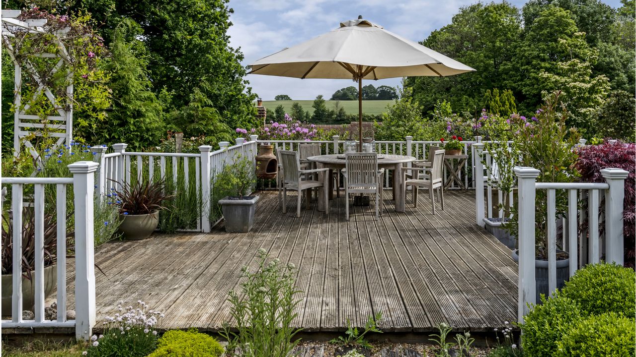 garden with wooden decking and pale railings with potted plants, outdoor table and chairs and parasol