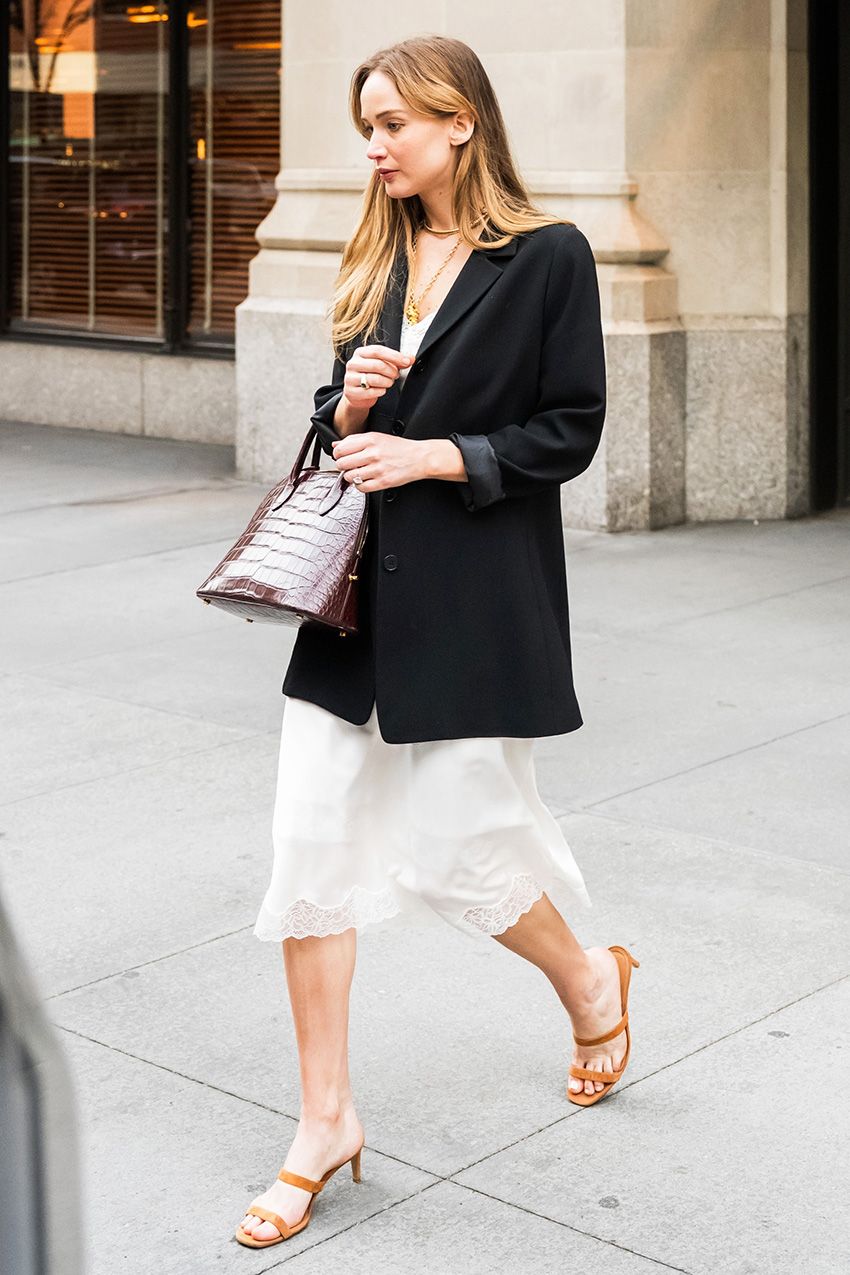 Jennifer Lawrence walking with her husband, wearing a black oversized blazer with a lace-trimmed white slip skirt, strappy nude high-heel sandals, and a croc The Row bag