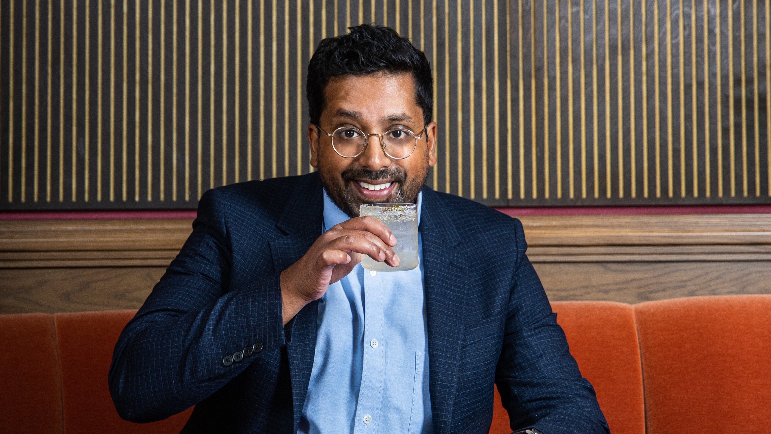 A young man with round silver glasses, a checkered blue tailored blazer, and light blue shirt sits on a velvety orange banquette before a paneled wood decorated in black and yellow stripes while holding a transparent cocktail.