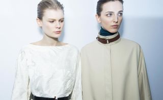 Two female models, one wearing a white top with belt, and one wearing a cream coat with brown belt at the neck