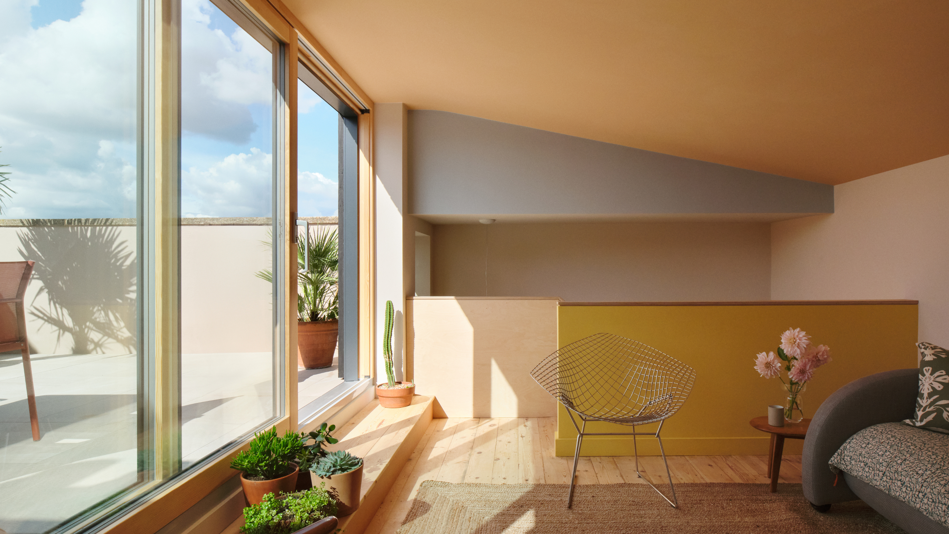An extension space with sloped ceiling painted in warm joyful tones with sliding doors, a step to the outside space with plants on it and a mesh metal chair next to a small table with flowers